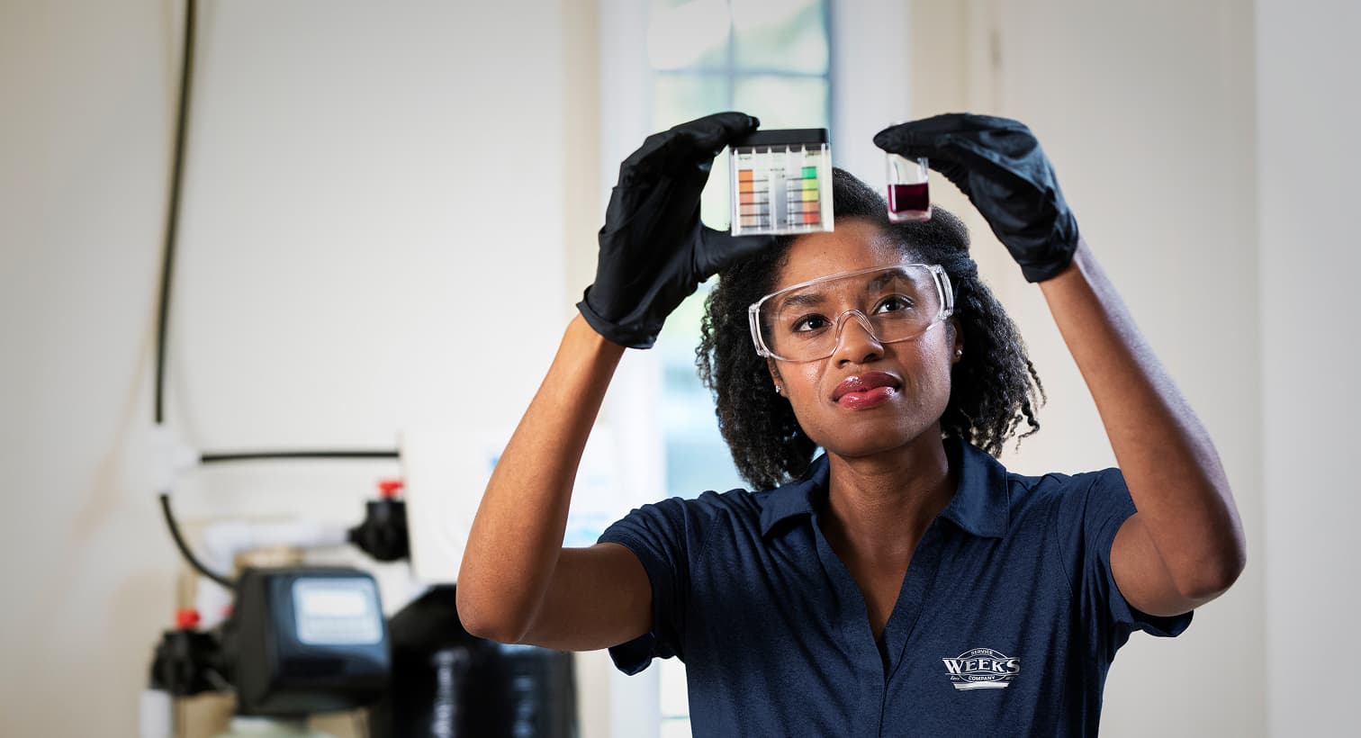 A Weeks technician in a blue uniform and safety gear compares a water sample to a testing chart in front of a water softener system.