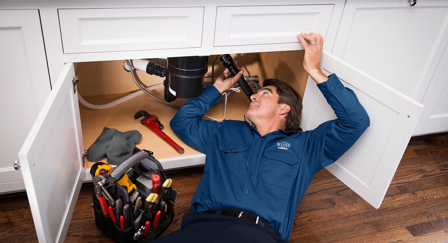 A professional Weeks Service Company plumber using a flashlight to inspect pipes under a kitchen sink for residential plumbing leak detection services.