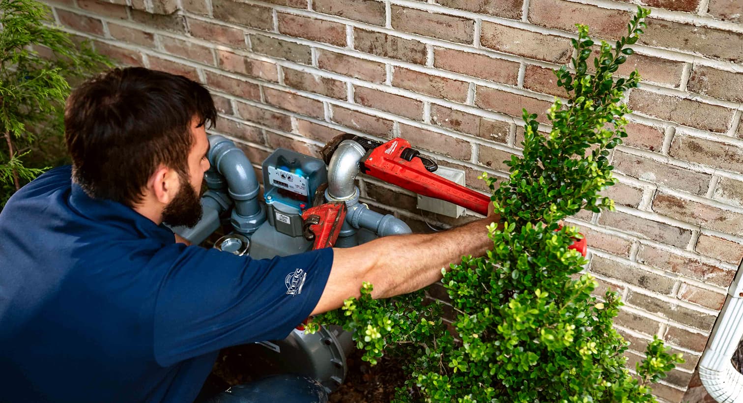 A professional Weeks Service Company technician using a red pipe wrench to service outdoor residential gas line plumbing and pipe connections against a brick wall.